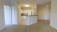 This single-floor residence features an open-concept layout that seamlessly connects the kitchen and living areas. The kitchen is outfitted with white cabinetry offering ample storage, dark countertops, and white appliances including a refrigerator and range. A built-in peninsula extends from the kitchen, providing additional counter space and serving as a natural divider between the kitchen and the adjacent living area. The floors transition from light tile in the kitchen to neutral-toned carpet in the living space, adding warmth and texture to the overall design. Crown molding accents the living area ceiling, lending a subtle architectural detail. The walls are finished in a soft beige tone throughout, contributing to a cohesive and understated aesthetic. A paneled door is visible near the kitchen, suggesting access to additional rooms or storage. The overhead fluorescent lighting in the kitchen ensures a well-lit workspace, while the open flow of the layout allows light to carry naturally into the living area, making the space feel airy and functional.