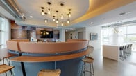 This thoughtfully designed single-floor commercial space blends contemporary aesthetics with functional versatility. At its centerpiece sits a bold circular bar or reception counter finished in warm caramel leather and rich wood tones, encircled by modern bar stools — an architectural focal point that immediately draws the eye. Above it, a curved tray ceiling with ambient uplighting and a dramatic cluster of industrial-style cage pendant lights adds depth and character to the space.

The open layout is defined by polished concrete flooring throughout, giving the environment a clean, urban feel. Floor-to-ceiling windows line the perimeter, flooding the interior with natural light and offering views of the surrounding cityscape. Sheer white curtains soften the light while maintaining an airy, open atmosphere.

A lounge area features a tan leather sofa, a velvet accent chair, a marble coffee table, and a textured feature wall with a wall-mounted flat-screen TV. A communal work table with built-in power access and leather seating sits beneath additional pendant lighting near a second mounted display. Glass-partitioned private offices provide focused work areas without sacrificing visual openness. A compact kitchenette with dark countertops, a beverage refrigerator, and a textured copper-toned accent wall completes the space. A long corridor connects additional rooms beyond the main area.