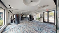 This well-appointed fitness center offers a thorough and thoughtfully organized workout environment across a single floor. The space is defined by its modern aesthetic, featuring crisp white walls, recessed lighting, and a series of circular ceiling cutouts housing ceiling fans that contribute to a clean, contemporary architectural character. A bold, multi-toned carpet in shades of blue, gray, and beige runs throughout, adding visual energy to the space.

Large windows line multiple walls, flooding the interior with natural light and providing views of the surrounding outdoor greenery. The equipment layout is both varied and generous. A dedicated cardio zone features a row of treadmills and ellipticals, each equipped with individual screens and positioned to face mounted televisions. A free weights area includes a full rack of dumbbells, adjustable benches, and a rowing machine. Strength training stations are distributed throughout, including cable machines and multi-functional resistance equipment.

A frosted-glass door leads to an accessible restroom, accompanied by a stainless steel water fountain nearby. Double glass entry doors at two points provide easy access to the outdoors. The overall ambiance is bright, clean, and functional, reflecting a facility designed for serious and casual exercisers alike.