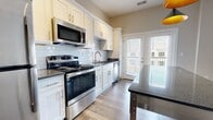 This contemporary apartment showcases a thoughtful design that maximizes space and functionality. The heart of the home is an open-concept kitchen, featuring crisp white cabinetry, stainless steel appliances, and a subway tile backsplash. A central island with a granite countertop provides additional workspace and casual dining options. The kitchen's modern pendant lighting adds a touch of elegance.

The living area is anchored by a sleek, built-in electric fireplace, creating a cozy atmosphere. Large windows and glass doors allow natural light to flood the space, while offering access to a private balcony. Rich, wood-look flooring unifies the entire apartment, adding warmth and durability.

The bathroom exudes luxury with its marble-inspired large format tiles, glass-enclosed shower, and modern vanity with ample storage. A barn door adds a rustic touch to the otherwise contemporary aesthetic. The bedroom, while not fully visible, appears spacious with the same high-quality flooring and features a sliding barn door for privacy.

The apartment's neutral color palette, combined with its high-end finishes and fixtures, creates a sophisticated and inviting living space perfect for modern urban living.