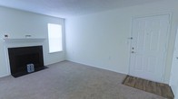 This single-floor residence offers a clean and functional layout designed for comfortable everyday living. The main living area is finished with wall-to-wall carpet in a neutral tone, creating a warm and inviting atmosphere throughout the space. Crisp white walls provide a bright, open feel, reflecting natural light that filters in through a window dressed with blinds. A classic fireplace with a dark surround serves as a notable focal point, adding both character and coziness to the room. The entryway features a solid white paneled door with brushed nickel hardware, transitioning from a hardwood-style entry mat into the carpeted living space — a practical and stylish detail. Smooth white baseboards frame the perimeter of the room, contributing to the clean, well-maintained aesthetic. The overall ambiance is understated and move-in ready, offering a neutral canvas that accommodates a wide variety of interior styles and furnishing choices. The single-floor design ensures ease of accessibility and a cohesive flow throughout the living space.