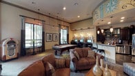 This spacious clubroom is thoughtfully designed to accommodate a variety of social and recreational activities within a single, open-plan layout. The space is anchored by a full-size billiards table positioned beneath a series of sleek pendant lights, flanked by large windows dressed with layered curtains that allow natural light to filter in. Bistro-style seating and framed artwork line the perimeter of this area, adding a refined touch.

A comfortable lounge area features plush, earth-toned sofas and accent chairs arranged around a coffee table, facing a wall-mounted flat-screen TV. The feature wall is uniquely designed with teal-painted recessed niches displaying decorative ceramics and art objects, creating a gallery-like focal point. A slatted wood partition subtly divides this zone from an adjacent seating area, maintaining an open yet defined flow.

A decorative rotunda with a painted mural ceiling adds architectural character and frames the transition toward the kitchenette, which includes a stainless steel refrigerator, bar seating, and pendant lighting over a granite-topped counter. A dedicated business center with computer workstations completes the space, making this clubroom a versatile and well-equipped communal hub.