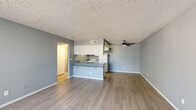 This efficient single-floor unit offers a well-organized layout comprising a combined kitchen and living room, one bedroom, one bathroom, and a hallway. The open-concept living and kitchen area is anchored by a peninsula-style counter with granite-look laminate surfaces, a stainless steel sink, white upper cabinetry, and white appliances including a refrigerator and range. Wood-look plank flooring runs throughout the main living area, complemented by neutral gray walls and a textured popcorn ceiling. A ceiling fan with light fixture adds functionality to the living space, which also features a large window allowing natural light to fill the room.

The bedroom is carpeted and includes a sliding-door closet for storage. A large window with vertical blinds allows ample natural light and outdoor views into the room. The bathroom is equipped with a full tub-shower combo, a toilet, and a granite-look countertop, finished with wood-look flooring consistent with the rest of the unit. Clean white walls and simple fixtures give the bathroom a straightforward, functional feel. Throughout the unit, a consistent cool gray color palette and clean white trim create a cohesive, modern aesthetic.