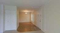 This single-floor space offers a clean, functional layout designed for comfortable everyday living. The entryway greets visitors with warm hardwood flooring that extends into the main living and dining area, providing a welcoming transition from the exterior. A classic white paneled front door with sidelights allows light to filter in, while a ceiling-mounted light fixture illuminates the space with a soft glow. Crown molding trims the ceiling throughout, adding a refined architectural detail to the otherwise understated interior. The neutral, warm beige walls create a bright and airy atmosphere that complements the natural tones of the flooring. A floor vent ensures efficient climate control throughout. Adjacent to the entry and dining area, the living space transitions to plush, light-toned carpeting, offering a softer underfoot experience. A large window dressed with vertical blinds dominates one wall, flooding the room with abundant natural light and providing an open, expansive feel. Baseboard heating runs along the perimeter, ensuring year-round comfort. The overall aesthetic is clean and neutral, making the space highly adaptable to a variety of furnishing styles and personal tastes.