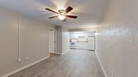 This single-floor unit is designed with a practical, open layout that connects the living area directly to the kitchen, creating a cohesive and functional living space. The main living area features light-toned luxury vinyl plank flooring and neutral gray walls, contributing to a bright, airy atmosphere. Ceiling fans with integrated light fixtures are present throughout, providing both ventilation and ambient lighting.

The kitchen is positioned at one end of the open living space and is equipped with white cabinetry, a dark granite-style countertop, a white electric range, an over-the-range microwave, and a full-size refrigerator. A skylight panel in the drop ceiling provides additional natural illumination to the kitchen area.

The bedroom is carpeted in a warm neutral tone and includes a ceiling fan, a window with blinds for natural light control, and access to the bathroom. A closet is accessible directly from the bedroom. The bathroom features a white vanity with under-cabinet storage, a large mirror with a multi-bulb light bar above, wood-look flooring, and brushed nickel hardware and fixtures, giving it a clean, updated appearance throughout.