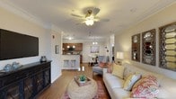 Located at 150 Horizon Trl in Garner, NC, this single-floor residence offers a thoughtfully designed open-concept layout that connects the main living spaces in a cohesive and functional flow. The living room serves as the heart of the home, featuring warm hardwood-style flooring, crown molding, and a ceiling fan with a light fixture that provides both comfort and illumination. The space is generously proportioned, allowing for comfortable furniture arrangements and easy movement throughout. A breakfast bar with barstool seating bridges the living area to the kitchen, which is outfitted with rich wood cabinetry, stainless steel appliances, and pendant lighting that adds a refined touch. Adjacent to the kitchen, the dining area accommodates a full dining set beneath an elegant chandelier, making it ideal for both casual meals and entertaining. Neutral, warm-toned walls throughout the home create a bright and inviting ambiance that complements the natural light filtering into the space. Crown molding and architectural columns add subtle elegance to the interior. A hallway visible from the main living area suggests additional private rooms beyond, completing this well-appointed, single-level home.