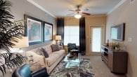 This single-floor residential space offers a thoughtfully designed living area that balances comfort and style. The main living room serves as the centerpiece of the home, featuring neutral-toned walls accented with crown molding that adds a refined architectural touch. A ceiling fan with integrated lighting fixture anchors the room, providing both airflow and ambient illumination.

Natural light fills the space through large, wide windows dressed with dark floor-length curtains, creating a dramatic contrast against the lighter walls. A glass side door with shuttered blinds provides additional brightness and access to the exterior. Soft wall-to-wall carpeting in a warm neutral tone adds comfort underfoot throughout the living area.

The room is thoughtfully arranged with a tufted cream sofa adorned with decorative pillows, positioned alongside a glass-top coffee table resting on a bold, patterned area rug. Bold abstract artwork in teal, blue, and grey tones adorns one wall, contributing a vibrant artistic focal point. A cozy seating nook near the window includes a leather armchair, making it an ideal spot for reading or relaxing.

A media console along one wall provides functional storage, while a table lamp adds warm, layered lighting. Overall, the space exudes a contemporary and inviting ambiance suited for comfortable everyday living.