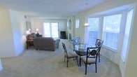 This single-floor residence offers an open-concept layout that seamlessly connects the living and dining areas, creating a spacious and cohesive environment. The dining area features a round glass-top table with elegant wrought-iron chairs, positioned to take full advantage of the abundant natural light streaming in through multiple large windows with white blinds. A pendant light fixture hangs overhead, adding a focused accent to the dining space.

The adjacent living area is generously sized, accommodating a plush, oversized sofa and coordinating seating arranged around a central fireplace — a defining architectural feature that serves as the room's focal point. A television and media console are neatly positioned nearby, enhancing the functionality of the space.

Throughout the open area, neutral-toned carpeting adds warmth and continuity, while the light-colored walls contribute to the airy, welcoming atmosphere. Sliding glass doors in the living area provide direct access to the outdoors, filling the space with additional natural light and offering views of the surrounding greenery. The overall ambiance is calm, comfortable, and well-suited for both everyday living and casual entertaining.