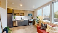 This thoughtfully designed single-floor residence combines modern aesthetics with functional living. The open-concept kitchen is a standout feature, showcasing two-tone cabinetry with warm wood uppers and slate-gray lower cabinets, complemented by a white geometric tile backsplash. Stainless steel appliances—including a gas range, built-in microwave, dishwasher, and refrigerator—are seamlessly integrated alongside a deep stainless farmhouse sink and clean white countertops. Open shelving adds a stylish, accessible storage element. The living area flows naturally from the kitchen, bathed in natural light from a large bank of windows that frame treetop views. Light wood-tone flooring runs throughout, unifying the space with warmth and continuity. The bedroom is a calm retreat with neutral white walls, a ceiling fan, and ample room for rest and relaxation. The bathroom features light wood vanity cabinetry, a tub-shower combination, black matte hardware accents, and light wood-tone flooring, creating a clean and contemporary feel. Recessed lighting throughout ensures each area is well-lit, while the generous window presence keeps the overall ambiance bright, airy, and connected to the outdoors.
