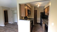 This single-floor unit offers a clean, functional layout designed for comfortable everyday living. The main living area is spacious and open, featuring rich, dark hardwood-style flooring that runs throughout the space. A large sliding glass door floods the room with natural light and provides direct access to a private balcony overlooking a green, tree-lined outdoor area — a standout feature that brings the outdoors in. Neutral warm-toned walls complement the flooring and create a cohesive, understated aesthetic throughout.

The kitchen is efficiently laid out and connects seamlessly to the living area via an open archway and a breakfast bar with dark countertops, making it ideal for casual dining or entertaining. Warm wood cabinetry lines the walls, providing ample storage, while black appliances including a built-in microwave and refrigerator add a modern contrast. Track lighting overhead ensures the kitchen is well-lit. A doorway off the kitchen leads further into the unit, offering additional private living spaces. A closet with shelving is also visible, providing practical storage solutions. Overall, the space presents a well-appointed, move-in ready environment with a balance of practicality and comfort.