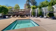 This outdoor pool area in Saugus, Massachusetts offers a thoughtfully designed recreational retreat. At its center is a generously sized swimming pool with crystal-clear turquoise water, accessible via stainless steel handrails and surrounded by a broad concrete sun deck ideal for lounging and socializing. Adjacent to the main pool is a separate hot tub, set against a striking natural stone retaining wall that adds a rugged, timeless character to the space.

The deck is well-furnished with modern grey chaise lounge chairs arranged throughout, including a dedicated sunbathing section shaded by a series of elegant white pergola structures. Tall columnar hedges and mature deciduous trees provide natural privacy screening and greenery throughout. Decorative planters with seasonal plantings are positioned thoughtfully across the patio, adding color and visual interest.

A classic shingle-style pool house with white trim anchors one end of the complex, providing utility access and storage. Additional amenities include patio umbrellas for shade and neatly trimmed hedgerows that define various zones within the space. The entire area is enclosed by black iron fencing and white vinyl gates, ensuring security while maintaining an open, airy atmosphere under abundant natural sunlight.