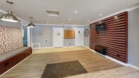 Located at 360 Sandra Ln in Indianapolis, IN, this single-floor community facility offers a thoughtfully designed layout encompassing a living room, laundry room, and exercise room. The welcoming living room serves as the central hub of the space, showcasing warm hardwood-style flooring, recessed lighting, crown molding, and a striking slatted wood accent wall fitted with a mounted TV and electric fireplace. Pendant lighting, a built-in bench with upholstered seating, and large windows allowing natural light complete the inviting atmosphere. Connecting rooms feature consistent gray paneled walls and light wood flooring throughout. The spacious laundry room is lined with a full row of commercial-grade front-load washers and dryers, complemented by a folding table, providing ample capacity for community use. The generously sized exercise room is divided into two well-lit areas: one equipped with stationary bikes, an elliptical, and a dumbbell rack with mirrored wall, and another dedicated to cardio with multiple treadmills and windows overlooking the outdoors. Textured ceilings and neutral gray tones carry throughout, giving the facility a clean, cohesive aesthetic that is both functional and comfortable.