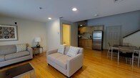 This two-floor residence offers a thoughtfully designed layout that balances comfort and functionality. The main living area is anchored by warm-toned hardwood floors that run throughout, lending a sense of continuity and natural warmth to the space. Recessed lighting overhead ensures bright, even illumination across the open floor plan.

The living area flows seamlessly into the kitchen, creating an open-concept feel ideal for both everyday living and entertaining. The kitchen is equipped with stainless steel appliances, including a full-size refrigerator, and features light wood cabinetry that adds a clean, understated aesthetic. A sink and countertop workspace complete the functional cooking area.

Neutral gray walls provide a versatile backdrop that complements the natural tones of the flooring and cabinetry. The layout makes efficient use of space while maintaining an open and airy atmosphere throughout the main level. A doorway visible from the living area hints at additional rooms beyond, contributing to the multi-room configuration spread across two floors.

Overall, the space presents a cohesive and practical interior with classic finishes, offering a comfortable environment with a clean, residential character throughout both levels.