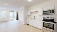 Located at 125 Liberty Ln in Nashville, TN, this modern 1-bedroom, 1-bathroom unit offers a thoughtfully designed open-concept layout finished with light hardwood flooring throughout. The combined kitchen and living area forms the heart of the unit, where a sleek kitchen features white shaker cabinetry, a stainless steel refrigerator with ice maker, electric range, built-in microwave, dishwasher, and a single-basin sink with a brushed nickel faucet. Clean white countertops and an open wood shelf accent the minimalist design. The living area flows naturally from the kitchen and opens to a private balcony via a sliding glass door framed by a dark curtain panel, offering views of the surrounding Nashville neighborhood. The bedroom is generously proportioned, filled with natural light from multiple windows with blinds, and also features direct balcony access through its own sliding glass door. A dedicated walk-in closet with wire shelving provides organized storage. The bathroom is equipped with a soaking tub and tiled surround, a white vanity with a rectangular mirror, and a small window for natural light. Recessed lighting, white panel doors with brushed nickel hardware, and crisp white walls complete the clean, contemporary aesthetic throughout.