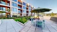Located at 377 Crane St in Orange, NJ, this thoughtfully designed outdoor amenity space serves as a central courtyard nestled within a contemporary multi-story residential building. The space is defined by a striking contrast between a vibrant green lawn and warm wood decking that spans the length of the courtyard, creating distinct zones for relaxation and social gathering.

Lush, manicured turf is flanked by carefully curated garden beds filled with ornamental grasses, flowering hydrangeas, and seasonal plantings, lending the space a park-like ambiance. Adirondack chairs positioned directly on the lawn invite casual outdoor lounging, while the expansive deck accommodates multiple seating configurations, including dining tables shaded by large green umbrellas and comfortable lounge furniture arranged for conversation.

The surrounding building facade features floor-to-ceiling windows, warm wood-accented balconies, and bold red architectural trim that adds visual energy to the space. The open-sky orientation ensures abundant natural light throughout the day. Large planter vessels punctuate the deck, adding greenery at varying heights and reinforcing the garden aesthetic. The overall ambiance is modern yet inviting, offering a well-appointed outdoor retreat designed for community living in an urban setting.