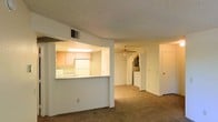 This single-floor residence offers a functional and open layout designed for comfortable everyday living. The living area is carpeted and features a textured popcorn ceiling, a ceiling fan for added air circulation, and a full-width sliding glass door dressed with vertical blinds that invite natural light to fill the room. The open design connects the living space seamlessly to the rest of the unit, creating a sense of continuity throughout.

The kitchen is accessed via a pass-through counter opening, which also serves as a breakfast bar. It is equipped with light wood cabinetry, a white refrigerator, a microwave, and overhead fluorescent lighting that brightens the workspace. The pass-through design keeps the kitchen visually connected to the main living area, making it well-suited for everyday use.

Beyond the main living space, an arched doorway leads further into the unit, where additional rooms are accessible. A washer unit is visible in a dedicated laundry area, adding in-unit convenience. Multiple interior doors suggest the presence of bedrooms and additional utility spaces. The overall interior palette is neutral, with white walls and warm-toned carpet, providing a clean and adaptable backdrop throughout the home.