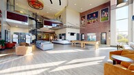 Located at 2525 W Tennessee St in Tallahassee, FL, this impressive clubhouse at Forum at Tallahassee offers a dynamic, open-concept layout spanning a generously sized single floor. The space is defined by dramatic double-height ceilings that amplify its airy, grand atmosphere, drawing in abundant natural light through expansive floor-to-ceiling windows that frame views of the surrounding greenery.

A striking black spiral staircase serves as a central architectural focal point, leading to an upper mezzanine level with glass railings that overlook the main floor below. Exposed brick walls adorned with large-scale sports-themed artwork add an energetic, collegiate character to the interior, complemented by warm wood-look tile flooring throughout.

The lounge areas are furnished with a mix of modular sofas, accent chairs, and coffee tables, creating comfortable and inviting gathering spaces. A well-appointed kitchen and bar area features navy cabinetry, white subway tile, stainless steel appliances, a white countertop island with bar seating, and a Starbucks coffee station. A shuffleboard table anchors the game zone, while multiple wall-mounted flat-screen TVs provide entertainment throughout. The entry foyer is welcoming and functional, completing this versatile and stylish amenity space.
