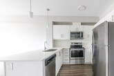 Located at 180 Greenbrier Rd in Cambridge, Ontario, this well-designed one-bedroom, one-bathroom unit offers a clean, modern aesthetic across a single floor. The open-concept living and kitchen area serves as the heart of the space, where a sleek peninsula with a double undermount sink and white quartz countertop connects the two zones seamlessly. The kitchen is outfitted with crisp white cabinetry, a white subway tile backsplash, and a full suite of stainless steel appliances including a refrigerator, electric range, built-in microwave, and dishwasher. Pendant lighting above the peninsula adds a contemporary touch. Light-toned laminate flooring runs through the main living area, transitioning to a darker wood-look finish in the kitchen, creating a subtle visual distinction between spaces. The living area benefits from a large window that brings in natural light and offers views of the surrounding neighbourhood. The bedroom is a comfortable, well-proportioned room featuring its own window with outdoor views and the same laminate flooring. A door leads to a private outdoor area. The bathroom includes a full soaker tub with overhead shower. In-suite laundry is also present, adding everyday convenience to this thoughtfully finished residence.