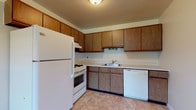 This single-floor residential unit offers a practical and comfortable layout across several well-defined living spaces. The kitchen is efficiently designed with warm oak cabinetry extending to the upper walls for ample storage, light-toned countertops, and tile flooring. It is fully equipped with a gas range, refrigerator, dishwasher, and a single-basin sink, making it a highly functional workspace. The kitchen opens toward the living area, which is finished with neutral wall-to-wall carpeting and features a ceiling fan with light fixture. Baseboard heating runs along the perimeter, and a window allows natural light to filter in. The transition between the kitchen's tile flooring and the living area's carpet marks a natural boundary between the two spaces. The bedroom is generously sized with the same plush carpeting, textured ceilings, and a notable double closet configuration featuring louvered bi-fold doors — offering substantial storage capacity. A window provides natural light and outdoor views. The bathroom is appointed with mosaic tile flooring in a classic pattern, white subway tile walls, a dark-countered vanity with under-sink cabinetry, a large wall mirror with vanity lighting, and a sliding glass door enclosure over the tub and shower. Overall, the unit presents a clean, neutral palette throughout.