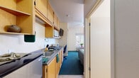 Located at 1485 Kerley Dr in San Jose, CA, this single-floor studio unit offers a thoughtfully organized layout that maximizes functionality within a compact footprint. The space is accessed through a secure entry door flanked by built-in open shelving, providing convenient storage from the moment you step inside. Adjacent to the entrance, a fully equipped kitchenette runs along one wall, outfitted with light wood cabinetry — both upper and lower — a stainless steel sink, microwave, toaster oven, and under-counter refrigerator, making everyday meal preparation straightforward and accessible. A wall-mounted flat-screen television is positioned above the kitchenette counter for easy viewing throughout the space. The living and sleeping area features rich teal carpeting and benefits from natural light through a window overlooking greenery. A ceiling fan keeps the space comfortable year-round, while a wall-mounted HVAC unit provides additional climate control. The sleeping area accommodates a full bed and includes a cozy seating corner with an upholstered armchair and side table. A built-in desk with additional cabinetry creates a defined workspace, complemented by an open closet with wire shelving for ample organization. The bathroom is finished with tile flooring and includes a shower-tub combination with sliding glass doors, a pedestal sink, and a wall mirror.