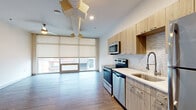This single-floor unit showcases a thoughtfully designed open-concept layout that seamlessly connects the living area and kitchen. The living space is defined by a dramatic floor-to-ceiling window wall fitted with roller shades, flooding the interior with natural light while offering views of the surrounding urban landscape. Wood-look plank flooring runs throughout, lending a warm, cohesive feel to the space, while a contemporary ceiling fan provides both function and a modern aesthetic touch.

The kitchen is well-appointed with light wood flat-front cabinetry, a textured tile backsplash, and white quartz countertops. A full suite of stainless steel appliances includes an electric range, over-the-range microwave, dishwasher, and refrigerator. A single-basin undermount sink with a sleek gooseneck faucet completes the workspace.

Beyond the main living area, a hallway leads to additional rooms, with double doors providing access to a generously sized closet or storage space. Louvered doors along the corridor suggest additional storage or utility areas. Clean white walls and recessed lighting throughout contribute to the unit's bright, contemporary ambiance, creating a move-in-ready space with a clean, modern aesthetic.