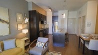 This single-floor space showcases an open-concept layout that seamlessly connects the kitchen and living area, creating a cohesive and functional environment. The kitchen is appointed with rich, dark espresso cabinetry that extends to the upper walls, providing ample storage. Stainless steel appliances, including a refrigerator and microwave, complement the mosaic tile backsplash that adds texture and visual interest. A central island with a granite or stone countertop serves as both a workspace and casual dining spot, flanked by bar-height stools. A pendant light hangs above the island, adding focused illumination alongside a track lighting system overhead. The hardwood flooring extends throughout, unifying the space with warmth and continuity. The living area features a neutral-toned armchair accented with decorative pillows, positioned atop a chevron-patterned area rug. Gray-toned walls provide a modern, urban backdrop, while the open sightlines between the kitchen and living zones enhance the overall sense of spaciousness. The layout is efficient yet inviting, balancing contemporary design elements with practical functionality.