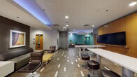 This contemporary single-floor commercial space is designed to offer both comfort and functionality in a polished, modern setting. The open layout flows seamlessly between distinct zones, each thoughtfully appointed with cohesive design elements. The main lounge area features plush upholstered sofas and seating arranged around low-profile side tables, set against warm charcoal-toned accent walls adorned with vibrant, colorful framed artwork. Soft wall sconces provide warm, ambient illumination that complements the recessed ceiling lighting throughout. A striking feature wall with textured paneling adds depth and visual interest to the space. Adjacent to the lounge, a sleek bar-height communal table with dark swivel stools is paired with wall-mounted flat-screen televisions and warm wood cabinetry, creating an ideal space for casual work or gathering. The patterned tile flooring — featuring a distinctive inlaid stripe motif — runs throughout the open area, tying the zones together with a clean, graphic element. Teal accent walls and ambient blue ceiling lighting add a bold pop of color near the central corridor, anchoring the space with a modern, energetic feel. A glass-walled fitness center is visible from the main area, offering views of exercise equipment within a bright, well-lit room. Elevators and restrooms are conveniently accessible, completing this well-rounded and thoughtfully designed communal environment.