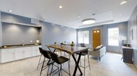 This clubroom offers a well-appointed communal space designed for both casual gatherings and productive meetups. The room is finished with soft blue-gray walls and light tile flooring, creating a clean, contemporary aesthetic throughout.

A striking live-edge wood communal table serves as the centerpiece, paired with sleek black bar-height chairs that provide seating for multiple guests. The natural wood grain of the table adds an organic warmth that contrasts effectively with the room's modern finishes.

Recessed lighting and a large circular flush-mount ceiling fixture ensure the space is well-lit, while a window allows natural light to filter in, adding to the open, airy feel of the room.

A built-in white cabinetry station along one wall provides a functional counter space, ideal for refreshments or casual use. Potted plants are thoughtfully placed throughout, contributing a touch of greenery to the interior.

A separate seating area near the entrance features a light-upholstered sofa and accent chair, offering a more relaxed lounge setting. The warm-toned wooden entry door adds a welcoming contrast to the cool color palette of the space.