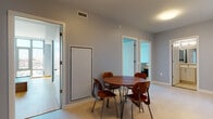 This single-floor residence is designed with a clean, contemporary aesthetic that is consistent throughout each room. The dining area serves as a central hub of the space, featuring light tile flooring, cool grey walls, and a mid-century modern round walnut dining table paired with molded wood chairs. Adjacent rooms are accessible through white-trimmed doorways, contributing to the open and connected feel of the layout. The kitchen is outfitted with stainless steel appliances — including a gas range, microwave, refrigerator, and dishwasher — complemented by crisp white cabinetry, a subway tile backsplash, and light stone countertops. The bedroom features warm hardwood flooring, a built-in wood headboard with integrated nightstands, dual closets with white panel doors, and direct access to the entry hallway. A standout feature is the private balcony, accessible through floor-to-ceiling glass doors, which frames expansive city views and fills the adjoining room with natural light. The space includes two bathrooms, each finished with light tile flooring, white vanities, and understated modern fixtures. Exposed concrete structural columns add an industrial design element that contrasts elegantly with the otherwise soft, neutral palette of the residence.
