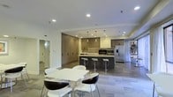 This single-level space showcases a contemporary open-plan layout that seamlessly integrates a fully equipped kitchen with a casual dining area. The kitchen is a standout feature, centered around a substantial dark island with built-in bar seating, accommodating multiple stools for casual dining or entertaining. Upper and lower cabinetry in warm wood tones lines the walls, complemented by a subtle tile backsplash and stainless steel appliances, including a refrigerator and built-in oven. Pendant lights hang above the island, adding a refined touch to the cooking space.

Adjacent to the kitchen, a dedicated dining area features a clean-lined white rectangular table paired with a mix of white and dark accent chairs, creating a striking visual contrast. The light wood-toned flooring runs throughout, contributing to a cohesive and airy atmosphere. Recessed ceiling lighting ensures the space is well-lit, while the open layout promotes a sense of spaciousness. A hallway visible in the background hints at additional rooms beyond. The overall design reflects a modern, functional aesthetic with attention to detail in materials and finishes.