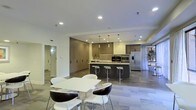 This single-level space showcases a contemporary open-plan layout that seamlessly integrates a fully equipped kitchen with a casual dining area. The kitchen is a standout feature, centered around a substantial dark island with built-in bar seating, accommodating multiple stools for casual dining or entertaining. Upper and lower cabinetry in warm wood tones lines the walls, complemented by a subtle tile backsplash and stainless steel appliances, including a refrigerator and built-in oven. Pendant lights hang above the island, adding a refined touch to the cooking space.

Adjacent to the kitchen, a dedicated dining area features a clean-lined white rectangular table paired with a mix of white and dark accent chairs, creating a striking visual contrast. The light wood-toned flooring runs throughout, contributing to a cohesive and airy atmosphere. Recessed ceiling lighting ensures the space is well-lit, while the open layout promotes a sense of spaciousness. A hallway visible in the background hints at additional rooms beyond. The overall design reflects a modern, functional aesthetic with attention to detail in materials and finishes.