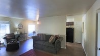 This single-floor residence offers a straightforward and functional layout designed for comfortable everyday living. The living area serves as the heart of the space, featuring warm hardwood-style flooring and neutral-toned walls that create a welcoming ambiance. A window allows natural light to fill the room, brightening the space and offering a view to the outside. The textured ceiling adds a subtle architectural detail overhead, while a ceiling fan fixture provides both comfort and casual illumination.

The open layout allows for a seamless transition from the living area toward the kitchen, which is visible from the main living space. The kitchen is equipped with dark cabinetry and a black refrigerator, offering a contrast against the lighter walls and floors. White interior doors contribute to the clean, cohesive aesthetic of the home.

The overall design is practical and unpretentious, with a layout that maximizes the use of available space. The combination of wood-look flooring, neutral walls, and natural light gives the residence a warm and inviting character that is both livable and easy to personalize.