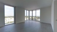 This sleek, contemporary space is defined by its striking floor-to-ceiling glass walls that wrap around a corner, flooding the interior with abundant natural light and offering expansive panoramic views of the surrounding cityscape and distant waterway. The open living area is finished with large-format grey tile flooring that lends a clean, refined aesthetic throughout. The minimalist design philosophy is evident in the smooth white walls and crisp white ceilings, creating a bright and airy ambiance that feels both spacious and modern. The corner window configuration maximizes the sense of openness, drawing the eye outward to sweeping vistas of treetops, urban rooftops, and a shimmering body of water on the horizon. A balcony is accessible through sliding glass doors, extending the living experience to the outdoors. The neutral color palette and understated finishes provide a versatile backdrop, allowing the breathtaking views to serve as the true centerpiece of the space. The overall atmosphere is one of elevated, contemporary urban living, with an emphasis on light, views, and clean architectural lines.