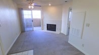 This single-floor residence offers a comfortable and functional layout centered around a welcoming living area. The room's most prominent feature is a classic tile-surround fireplace, framed in white ceramic tile, which serves as a natural focal point and adds warmth to the space. Plush wall-to-wall carpeting extends throughout the living area, contributing to a cozy ambiance underfoot.

Natural light flows generously into the space through a large sliding glass door, which opens to an outdoor area — ideal for extending living and entertaining space. A ceiling fan with an integrated light fixture is centrally positioned overhead, providing both air circulation and supplemental lighting. Additional recessed lighting in the ceiling further brightens the interior.

The flooring transitions from carpet to tile at the entryway, hinting at a thoughtful separation of zones within the home. A secondary window on the adjacent wall allows for cross-ventilation and added daylight. An HVAC vent is integrated into the wall, and a doorway visible from the living area connects to the rest of the home's interior. Overall, the space presents a clean, neutral palette with practical features throughout.