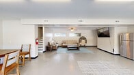 Located at 2215 Montauk Rd in Roanoke, Virginia, this single-floor gathering space offers a well-organized and functional layout designed for comfortable communal use. The interior is divided into two distinct areas, each serving a clear purpose while maintaining an open, airy feel throughout.

The lounge area features polished concrete floors, a ceiling fan, and horizontal windows that draw in natural light. It is furnished with wicker sofas, a hanging egg chair, a wall-mounted flat-screen TV, and a stainless steel refrigerator, creating a relaxed and welcoming atmosphere. A staircase along one wall provides additional access points within the building.

Adjacent to the lounge is a bright dining and gathering area, highlighted by large windows and glass French doors that open directly to an outdoor patio. A central dining table with wooden chairs anchors the space, while a large abstract artwork adds a decorative focal point. Fluorescent ceiling fixtures ensure consistent, even lighting throughout.

A compact kitchenette rounds out the space, equipped with white cabinetry, granite-style countertops, a stainless steel microwave, and a bulletin board display area near a large window overlooking the exterior. The neutral color palette, polished floors, and clean lines throughout give the space a practical yet inviting character, well-suited for meetings, events, or everyday community gatherings.