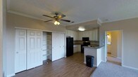 This single-floor space showcases an open-concept layout that seamlessly connects the living area and kitchen, creating a functional and cohesive environment. The flooring throughout the main area consists of warm hardwood-style planks, transitioning to carpet in an adjacent room visible in the background.

The kitchen is well-appointed with white raised-panel cabinetry offering generous storage. Black appliances, including a refrigerator, range, and over-the-range microwave, provide a classic contrast against the light cabinetry. A peninsula with a dark countertop extends from the kitchen, serving as a natural divider between the cooking and living spaces while offering additional workspace.

Crown molding runs along the upper perimeter of the room, adding an elegant architectural detail. The textured ceiling features a ceiling fan with an integrated light fixture, providing both air circulation and ambient lighting. Built-in shelving is integrated into the wall alongside a set of bifold closet doors, offering practical storage solutions. The overall ambiance is neutral and inviting, with a warm, understated palette throughout.