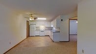 This single-floor space features an open layout that connects the kitchen and living area, creating a functional and cohesive environment. The kitchen is outfitted with white cabinetry that extends to the upper and lower sections of the walls, providing ample storage. A double-basin sink is positioned along the countertop, which runs in an L-shaped configuration to maximize workspace. White appliances, including a refrigerator, electric range with oven, and what appears to be a dishwasher, are integrated seamlessly into the kitchen design. An under-cabinet range hood is installed above the cooktop for ventilation. The flooring in the kitchen area features a light-toned tile pattern, while the adjoining living space transitions to carpeting, adding warmth underfoot. A ceiling fan with a built-in light fixture is centrally mounted, providing both air circulation and ambient lighting throughout the open area. The walls are finished in a neutral tone, contributing to a clean and simple aesthetic. A doorway visible in the background suggests additional rooms or access points beyond the main living space, while another doorway opens to a separate room with matching light-toned carpeting.