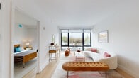 Located at 15 Bond Street in Great Neck Plaza, NY, this contemporary residence offers a thoughtfully designed single-floor layout with clean lines and a bright, modern aesthetic. The open-plan living and dining area is a standout feature, anchored by floor-to-ceiling windows with bold black frames that flood the space with natural light and provide views of the surrounding neighborhood. Light hardwood flooring runs throughout, complementing the crisp white walls and creating a warm, cohesive atmosphere. The kitchen is seamlessly integrated into the living area, centered around a generous island with a white countertop, undermount sink, and warm wood cabinetry. Stainless steel appliances, including a range and refrigerator, complete the functional and stylish cooking space. A dedicated dining area with a round table sits adjacent to the kitchen, ideal for entertaining. The residence includes two bedrooms, each well-proportioned with large windows, soft neutral tones, and ample natural light. The bathrooms feature a consistent design language with light wood vanities, white countertops, large framed mirrors, and grey tile flooring. The primary bathroom includes a glass-enclosed walk-in shower. Overall, the space exudes a refined, contemporary character with high-quality finishes and a well-considered layout.