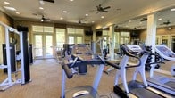 This fitness center offers a thoughtfully designed, single-level space dedicated to health and wellness. The room is outfitted with a variety of cardio machines, including treadmills and ellipticals from brands such as Spirit, alongside strength training equipment to support a full-body workout routine. Large floor-to-ceiling windows and glass-paneled doors line one wall, flooding the space with natural light and providing refreshing views of the surrounding outdoor greenery. Multiple ceiling fans are distributed evenly throughout the room, ensuring consistent air circulation during workouts. Recessed lighting complements the natural light, keeping the space bright and well-lit at all times. A wall of mirrors enhances the sense of openness and allows users to monitor their form while exercising. Wall-mounted televisions are positioned for easy viewing from the equipment. The flooring features low-pile carpet, providing comfort underfoot while maintaining a functional workout environment. Decorative stone or tile accents on the walls contribute a warm, upscale aesthetic that elevates the overall ambiance of the facility. The combination of practical amenities, thoughtful design, and an airy atmosphere makes this fitness center a welcoming and fully functional space for daily exercise.