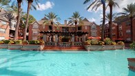 Located at 4848 Pin Oak Park in Houston, Texas, this single-floor residence offers a well-appointed living experience within a beautifully landscaped community. The exterior impresses immediately, showcasing classic brick architecture set against lush tropical palm trees and a stunning resort-style pool complete with water features, a pergola-covered lounge area, and vibrant outdoor seating — an inviting retreat right at your doorstep.

Inside, the entryway greets residents with clean, neutral tones and tile flooring, providing a practical and welcoming transition into the home. Rich, dark hardwood-style flooring flows throughout the main living areas, lending warmth and a sense of continuity. The living room is spacious and open, featuring a ceiling fan, soft neutral walls, and ample room for comfortable furnishings.

A hallway with pendant lighting and a curated gallery wall adds artistic character, while louvered closet doors provide functional storage. The kitchen and bathroom areas feature dark cabinetry with granite-style countertops, offering a sleek, contemporary aesthetic. Neutral gray walls and crisp white trim are consistent throughout, creating a cohesive and modern interior palette. The overall ambiance is calm, polished, and move-in ready.