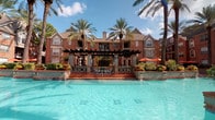 Located at 4848 Pin Oak Park in Houston, Texas, this single-floor residence offers a well-appointed living experience within a beautifully landscaped community. The exterior impresses immediately, showcasing classic brick architecture set against lush tropical palm trees and a stunning resort-style pool complete with water features, a pergola-covered lounge area, and vibrant outdoor seating — an inviting retreat right at your doorstep.

Inside, the entryway greets residents with clean, neutral tones and tile flooring, providing a practical and welcoming transition into the home. Rich, dark hardwood-style flooring flows throughout the main living areas, lending warmth and a sense of continuity. The living room is spacious and open, featuring a ceiling fan, soft neutral walls, and ample room for comfortable furnishings.

A hallway with pendant lighting and a curated gallery wall adds artistic character, while louvered closet doors provide functional storage. The kitchen and bathroom areas feature dark cabinetry with granite-style countertops, offering a sleek, contemporary aesthetic. Neutral gray walls and crisp white trim are consistent throughout, creating a cohesive and modern interior palette. The overall ambiance is calm, polished, and move-in ready.