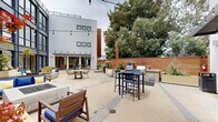 Located at 555 Pacific Ave in Santa Cruz, California, this striking outdoor courtyard space showcases a thoughtfully designed communal environment framed by a soaring multi-story building façade dominated by expansive floor-to-ceiling steel-framed windows. The towering glazed wall floods the courtyard with natural light and creates a seamless visual connection between the interior and exterior.

The central courtyard is generously proportioned and finished with polished concrete flooring. It features multiple distinct seating areas, including a built-in fire pit surrounded by wood-framed chairs and sofas upholstered in bold cobalt blue, complemented by accent pillows in yellow and white. Large planters with lush greenery are positioned throughout, adding warmth and a natural element to the otherwise modern aesthetic. String lights overhead lend the space an inviting ambiance for evening gatherings.

A dedicated outdoor kitchen area lines one edge of the courtyard, equipped with a gas grill, pizza oven, and built-in cabinetry — ideal for entertaining. Adjacent to the kitchen, casual bar-height seating and a round dining table with woven chairs provide additional gathering options. The courtyard is enclosed by a warm cedar privacy fence and mature trees, creating a sheltered and tranquil atmosphere within an urban setting.