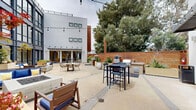 Located at 555 Pacific Ave in Santa Cruz, California, this striking outdoor courtyard space showcases a thoughtfully designed communal environment framed by a soaring multi-story building façade dominated by expansive floor-to-ceiling steel-framed windows. The towering glazed wall floods the courtyard with natural light and creates a seamless visual connection between the interior and exterior.

The central courtyard is generously proportioned and finished with polished concrete flooring. It features multiple distinct seating areas, including a built-in fire pit surrounded by wood-framed chairs and sofas upholstered in bold cobalt blue, complemented by accent pillows in yellow and white. Large planters with lush greenery are positioned throughout, adding warmth and a natural element to the otherwise modern aesthetic. String lights overhead lend the space an inviting ambiance for evening gatherings.

A dedicated outdoor kitchen area lines one edge of the courtyard, equipped with a gas grill, pizza oven, and built-in cabinetry — ideal for entertaining. Adjacent to the kitchen, casual bar-height seating and a round dining table with woven chairs provide additional gathering options. The courtyard is enclosed by a warm cedar privacy fence and mature trees, creating a sheltered and tranquil atmosphere within an urban setting.