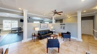 Located at Tripp Cottages in Siler City, North Carolina, this single-floor community space offers a thoughtfully designed layout that serves both social and functional needs. The living area greets visitors with warm, neutral-toned walls accented by crisp white crown molding, decorative columns, and tile flooring throughout. Ceiling fans and recessed lighting provide comfort and brightness, while the open layout fosters a welcoming atmosphere. Comfortable seating arrangements, including a leather sofa and armchairs, make this an ideal gathering space, and double doors allow for natural light and easy access. The kitchen is generously sized and well-equipped, featuring warm wood cabinetry, granite-style countertops, a central island with bar seating, and black appliances including a refrigerator and microwave. A dedicated sink area with ample counter space and extensive cabinet storage make this kitchen fully functional for community events and everyday use. The vaulted ceiling in the kitchen adds an open, airy feel to the space. An adjoining fitness area rounds out the amenities, housing a variety of exercise machines to support an active lifestyle. Together, these spaces create a versatile, community-focused environment designed for comfort, convenience, and connection.