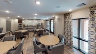 This single-floor communal space is thoughtfully designed to serve as both a social gathering area and a fully functional kitchen and dining facility. The kitchen is appointed with rich dark cabinetry, granite countertops, stainless steel appliances including a wall oven, microwave, and refrigerator, and a striking textured tile backsplash that serves as a focal point. A coffee and beverage station adds convenience along the extended counter run. The flooring throughout features wood-look planks laid in a herringbone-style pattern, lending warmth and visual depth to the space. The dining area accommodates multiple round tables surrounded by upholstered nailhead-trim chairs, creating an inviting atmosphere for group gatherings or casual meals. Bold patterned drapery panels frame large windows and glass doors that open to an exterior balcony, allowing natural light to filter through while maintaining a sense of privacy. A separate lounge zone offers plush seating arrangements with accent stools and wall art, set beneath a dramatic dark-painted tray ceiling with pendant lighting. Recessed lighting throughout ensures consistent illumination, while the cohesive neutral color palette with navy blue accents ties all areas of the space together into a polished, contemporary aesthetic.
