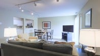 This two-floor residence is designed with an open-concept layout on the main level, creating a cohesive flow between the living, dining, and kitchen areas. The living room is generously sized, anchored by a plush sectional sofa and a comfortable armchair, with a flat-screen television mounted on a media console positioned near a bright window that allows natural light to fill the space. Track lighting runs along the ceiling, providing adjustable illumination throughout the room.

The dining area sits adjacent to the living space, featuring a classic wooden table with matching chairs, making it well-suited for everyday meals or gatherings. A framed piece of artwork adds a decorative touch to the shared wall between these zones.

The kitchen is partially open to the main living area via a pass-through opening, offering visibility and connectivity. It is equipped with stainless steel appliances, including a microwave, and features light cabinetry. The overall color palette throughout the main level is soft and neutral, with light walls and carpeted flooring contributing to a calm, welcoming ambiance. The second floor provides additional living space, expanding the home's overall functionality.