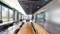 Located at 1950 Broadway in Oakland, CA, this single-floor amenity lounge is a thoughtfully designed social space that blends modern industrial aesthetics with refined, upscale finishes. The open-plan layout is anchored by wide-plank hardwood flooring, dramatic black slatted ceiling beams, and expansive floor-to-ceiling glass windows that flood the interior with natural light and frame views of the surrounding urban landscape and greenery.

A striking dark granite-clad island and a backlit bar wall featuring dramatic veined stone serve as the room's centerpiece, creating an elegant focal point for entertaining. A long communal wood dining table with gold-accented candleholders and curved upholstered seating provides a central gathering space, while multiple lounge vignettes — including leather and fabric chairs with brass-finished legs — offer versatile seating arrangements throughout.

A foosball table adds a recreational dimension to the space, complementing the room's social character. Pendant lighting with a brushed metallic finish descends gracefully above key seating areas, enhancing the warm, sophisticated ambiance. Textured wood-panel walls and frosted glass partitions contribute refined architectural layering. The overall atmosphere is polished yet inviting, making this lounge an exceptional communal retreat.