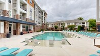 Located at 2301 E Tenth St in Greenville, North Carolina, this outdoor amenity area serves as a central gathering space within a well-appointed residential community. The centerpiece is a generously sized resort-style swimming pool with clear, blue-green water, designed to accommodate both leisurely lounging and active recreation. A dedicated wading shelf along one side of the pool offers a shallow, sun-soaked area ideal for relaxing directly in the water. The expansive pool deck is finished in smooth concrete and lined with an abundance of white and teal-cushioned lounge chairs, providing ample space for sunbathing and outdoor relaxation. The pool area is enclosed by a black wrought iron fence, enhancing both security and aesthetic appeal. Mature evergreen shrubs, ornamental trees, and seasonal plantings are thoughtfully integrated along the perimeter, adding lush greenery to the hardscape. A covered outdoor lounge area with seating is positioned adjacent to the pool, offering a shaded retreat. Beyond the pool deck, a dedicated courtyard space with turf and open concrete provides additional outdoor recreation opportunities. The multi-story buildings surrounding the area feature a combination of brick, siding, and wood accents with private balconies, framing the outdoor space and contributing to an active, community-oriented atmosphere.