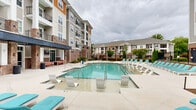 Located at 2301 E Tenth St in Greenville, North Carolina, this outdoor amenity area serves as a central gathering space within a well-appointed residential community. The centerpiece is a generously sized resort-style swimming pool with clear, blue-green water, designed to accommodate both leisurely lounging and active recreation. A dedicated wading shelf along one side of the pool offers a shallow, sun-soaked area ideal for relaxing directly in the water. The expansive pool deck is finished in smooth concrete and lined with an abundance of white and teal-cushioned lounge chairs, providing ample space for sunbathing and outdoor relaxation. The pool area is enclosed by a black wrought iron fence, enhancing both security and aesthetic appeal. Mature evergreen shrubs, ornamental trees, and seasonal plantings are thoughtfully integrated along the perimeter, adding lush greenery to the hardscape. A covered outdoor lounge area with seating is positioned adjacent to the pool, offering a shaded retreat. Beyond the pool deck, a dedicated courtyard space with turf and open concrete provides additional outdoor recreation opportunities. The multi-story buildings surrounding the area feature a combination of brick, siding, and wood accents with private balconies, framing the outdoor space and contributing to an active, community-oriented atmosphere.