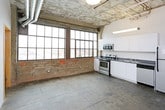 This single-floor urban loft masterfully blends industrial heritage with modern living. The open layout is defined by striking polished concrete floors that run throughout, exposed concrete ceilings with visible ductwork and conduit, and original brick accent walls that speak to the building's storied past. Oversized, multi-pane industrial windows flood the living and kitchen areas with natural light while offering expansive views of the surrounding cityscape.

The kitchen is well-appointed with white cabinetry, dark countertops, and a full suite of stainless steel appliances including a range, microwave, dishwasher, and refrigerator. The open kitchen flows into a generous living area, making the space ideal for both daily living and entertaining.

The sleeping quarters feature sliding wood-panel closet doors and clean white walls that provide a calm contrast to the raw industrial elements found elsewhere. Exposed brick columns and high ceilings carry through, maintaining the loft's architectural character.

Two bathrooms serve the space, each featuring a walk-in shower with white surrounds, a toilet, and polished fixtures. One bathroom includes a vanity with a vessel sink and mirror. Throughout the unit, the interplay of raw materials and modern finishes creates a dynamic, warehouse-inspired ambiance.
